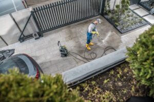 Professional maintaining a concrete driveway, cleaning and sealing cracks in front of a suburban house.